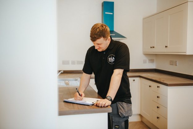 An electrician stood in a kitchen, leaning on the counter top and writing on a checklist.